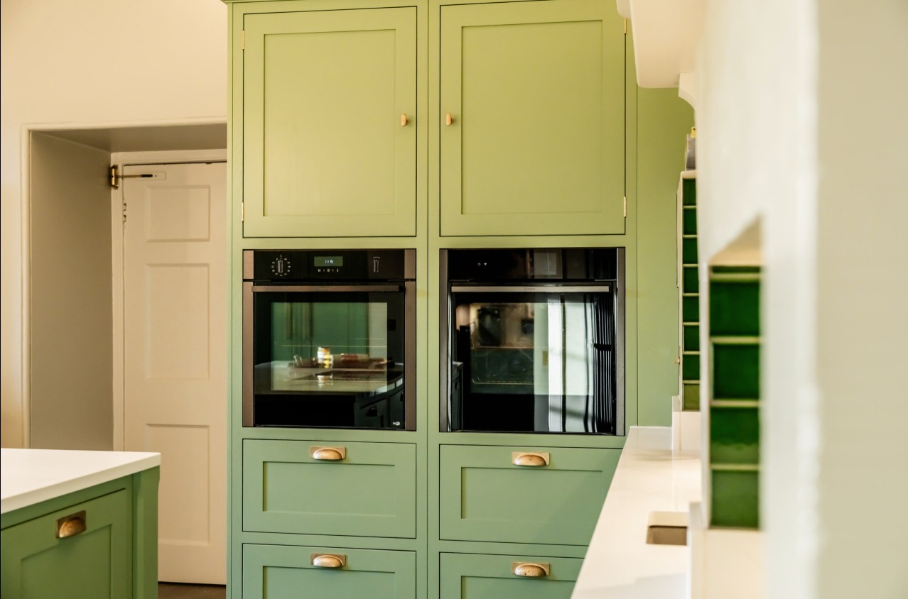 Green shaker kitchen with integrated ovens, brass cup handles and green tile splashback by 21 Kitchens in Ayrshire.