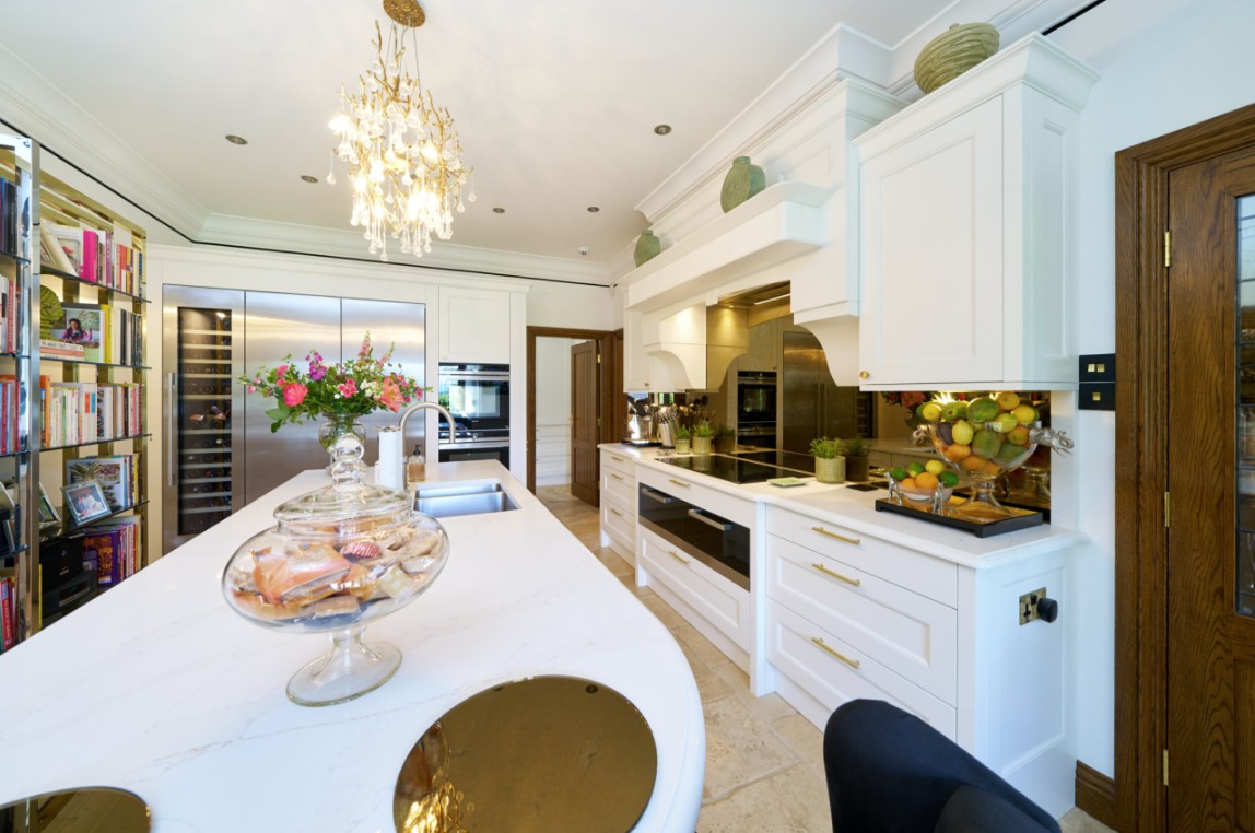 Luxury white shaker kitchen with a large quartz island, built in wine fridge, integrated appliances, brass hardware, crystal chandelier, limestone flooring, and fresh floral arrangements in a bright, elegant space.