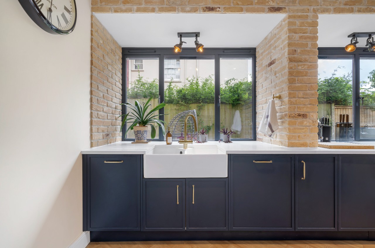 Belfast sink with brass tap in a contemporary Nobilia kitchen near Stewarton, Ayrshire, featuring navy cabinetry, white quartz worktops and exposed brick walls.