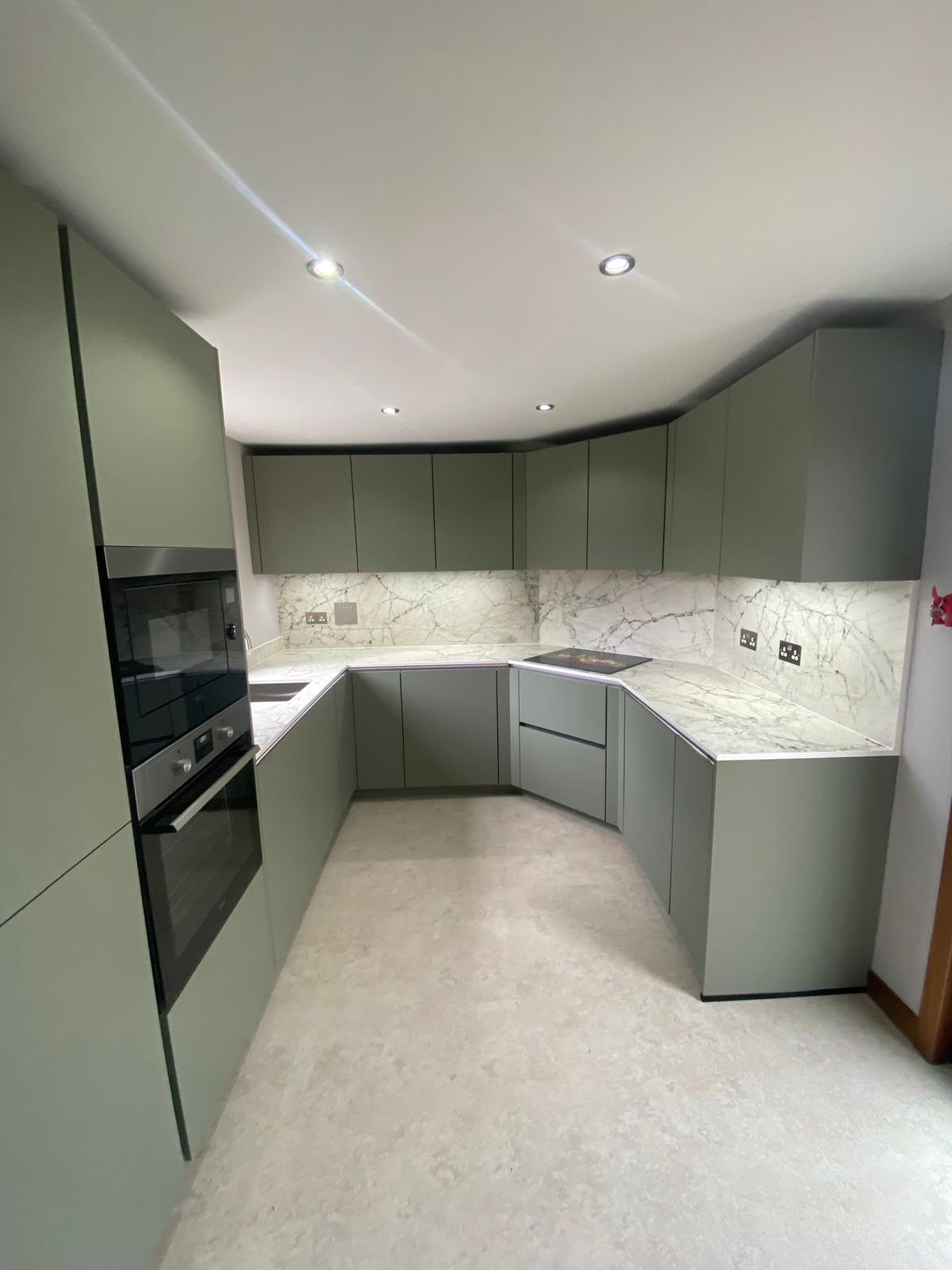 Sage green U-shaped kitchen with built-in oven, marble-effect worktops, sink and under-cabinet lighting.