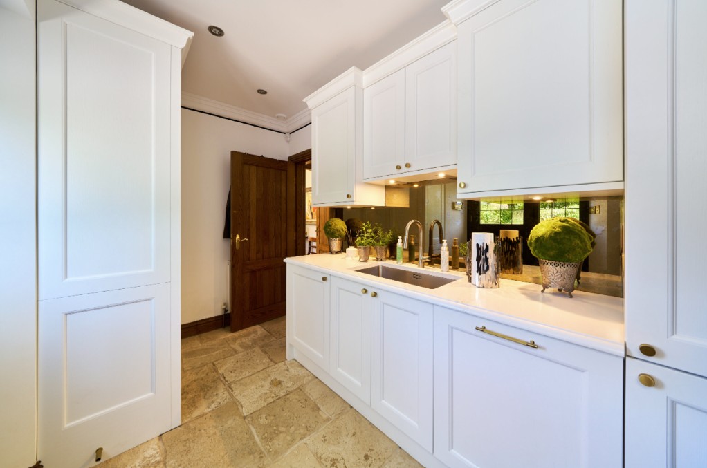 White shaker kitchen preparation area with integrated sink, quartz worktop, brass hardware, mirrored splashback, full height storage cabinets, under cabinet lighting, and natural limestone flooring.